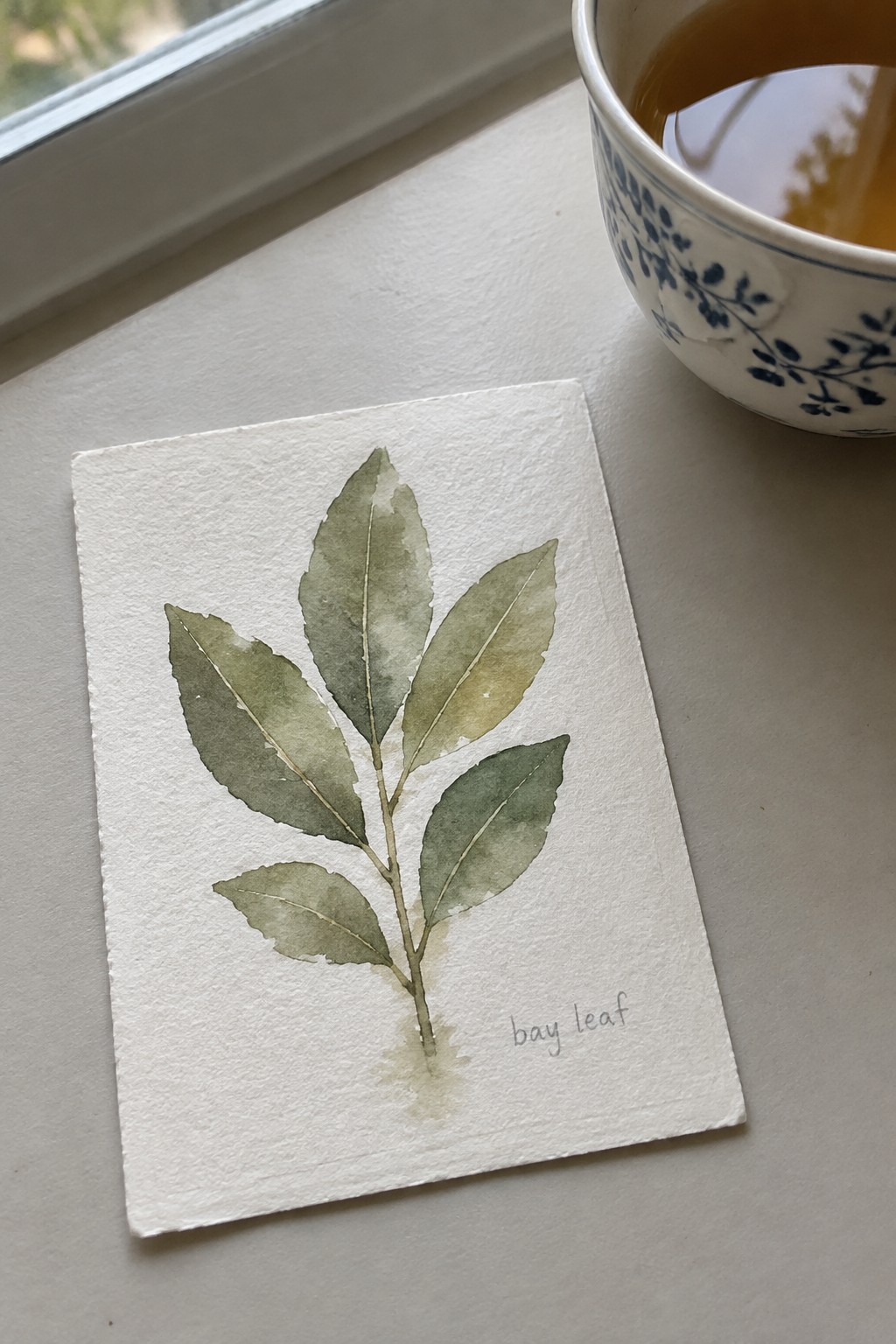 Watercolor bay leaf painting on paper beside blue floral teacup on windowsill.