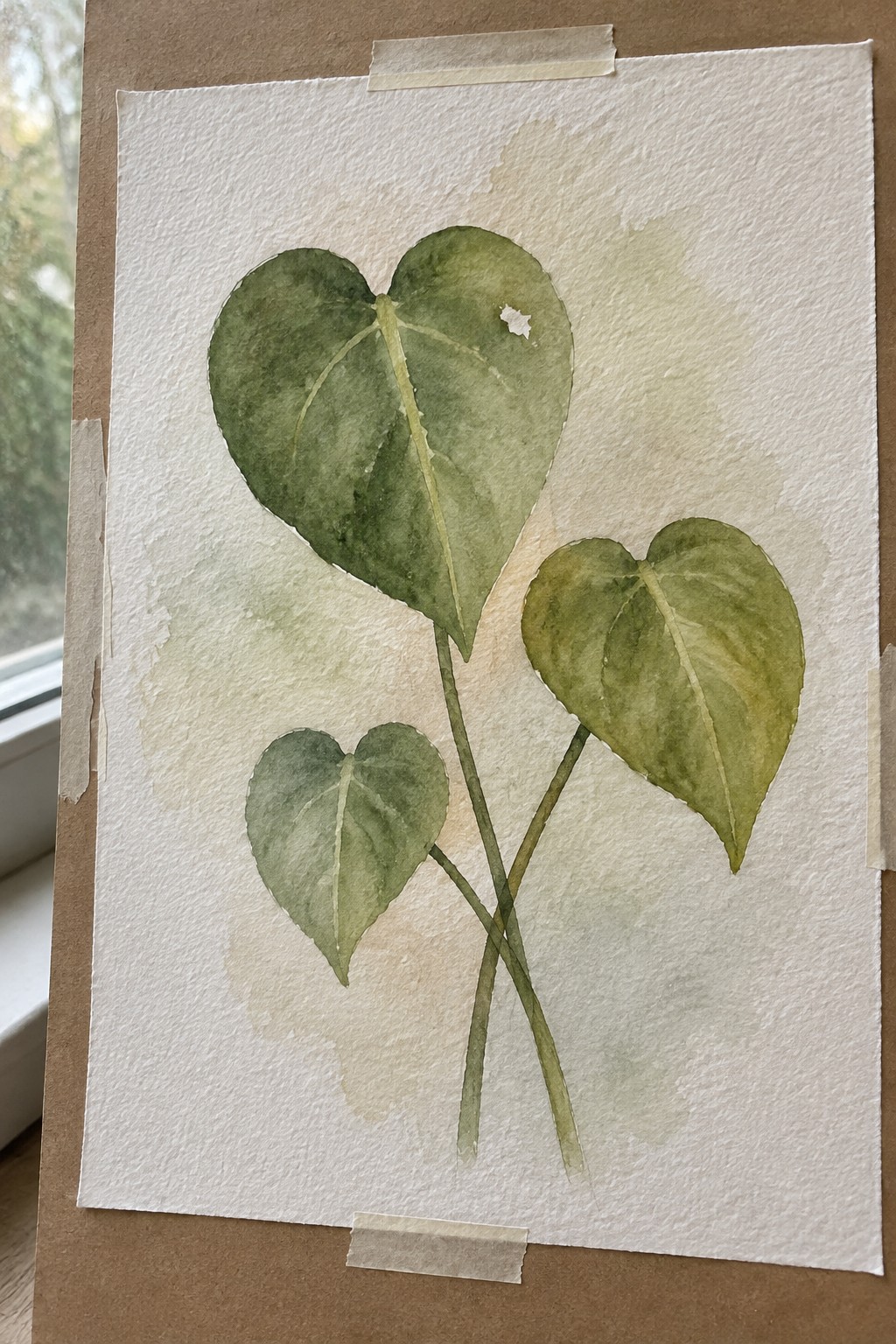 Three green watercolor leaves on crossed stems, taped to white paper on brown board.
