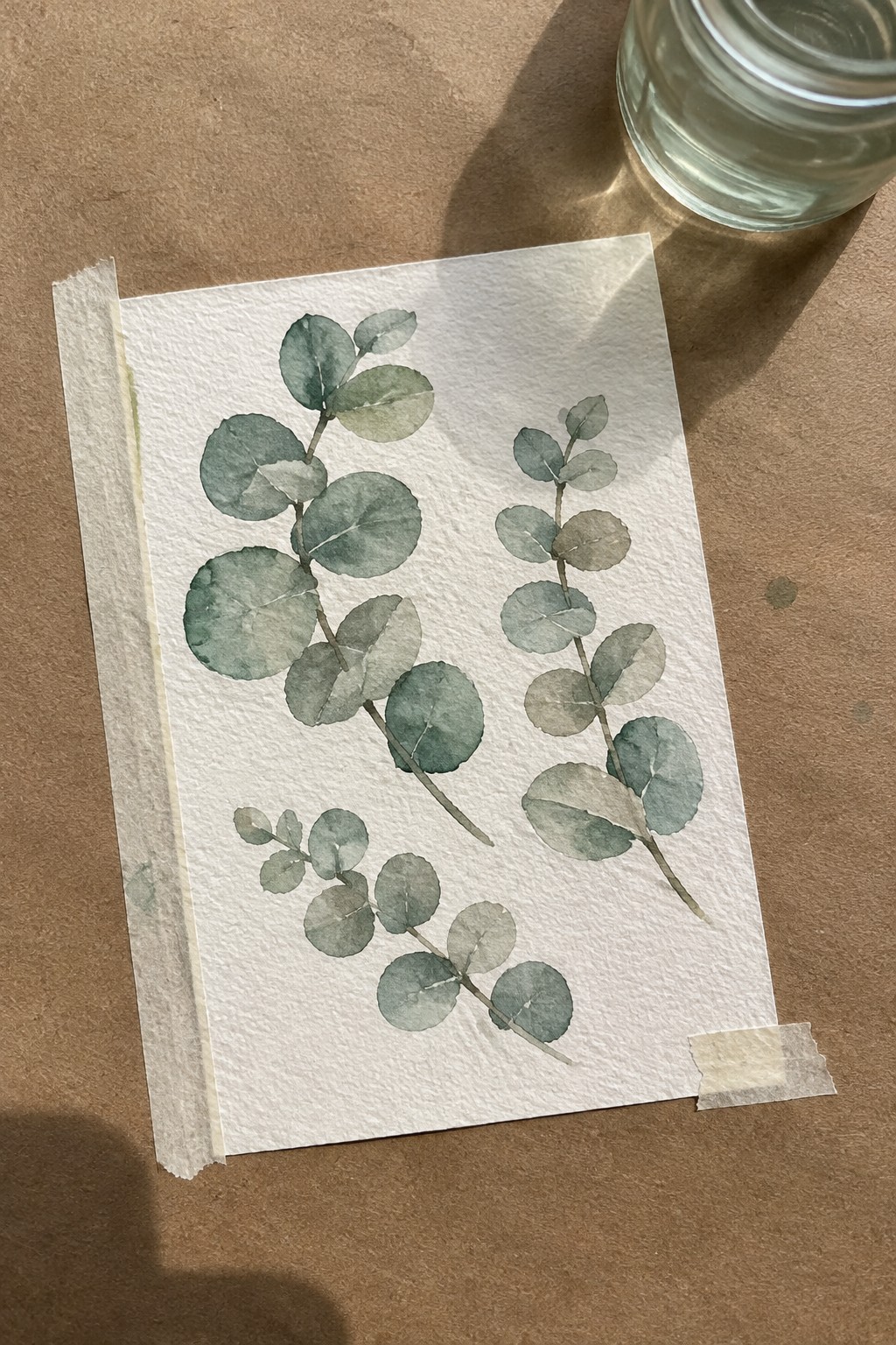 Watercolor eucalyptus branches in green tones on taped white paper beside glass jar on brown surface