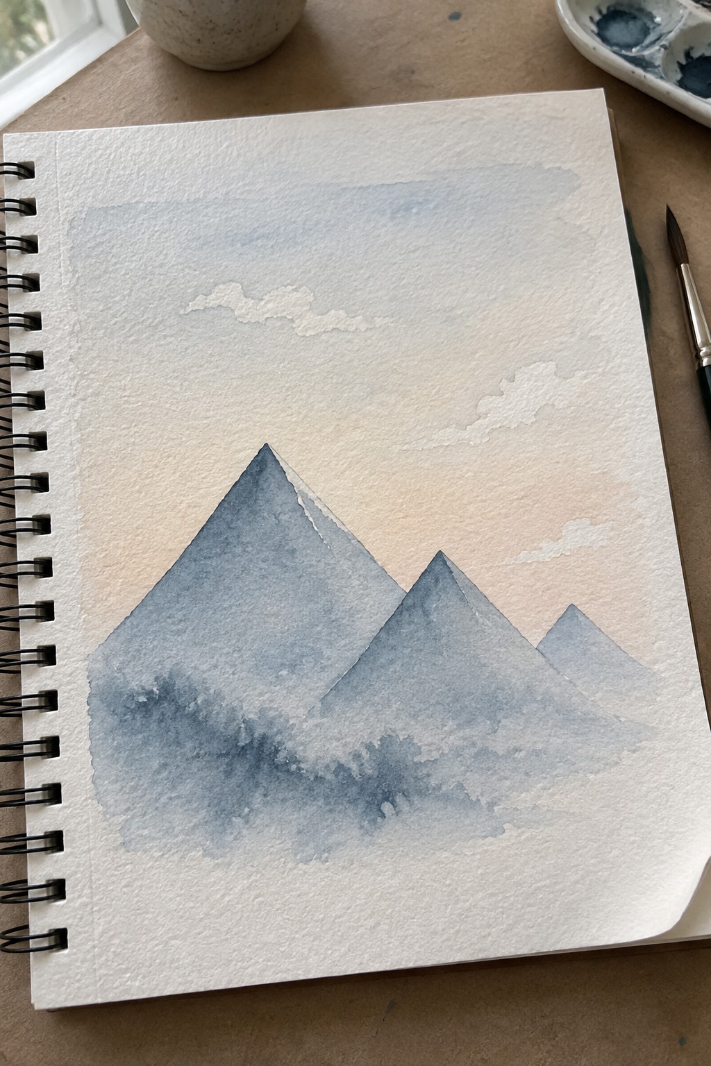 Watercolor painting of three blue mountains in open spiral sketchbook beside paintbrush and palette
