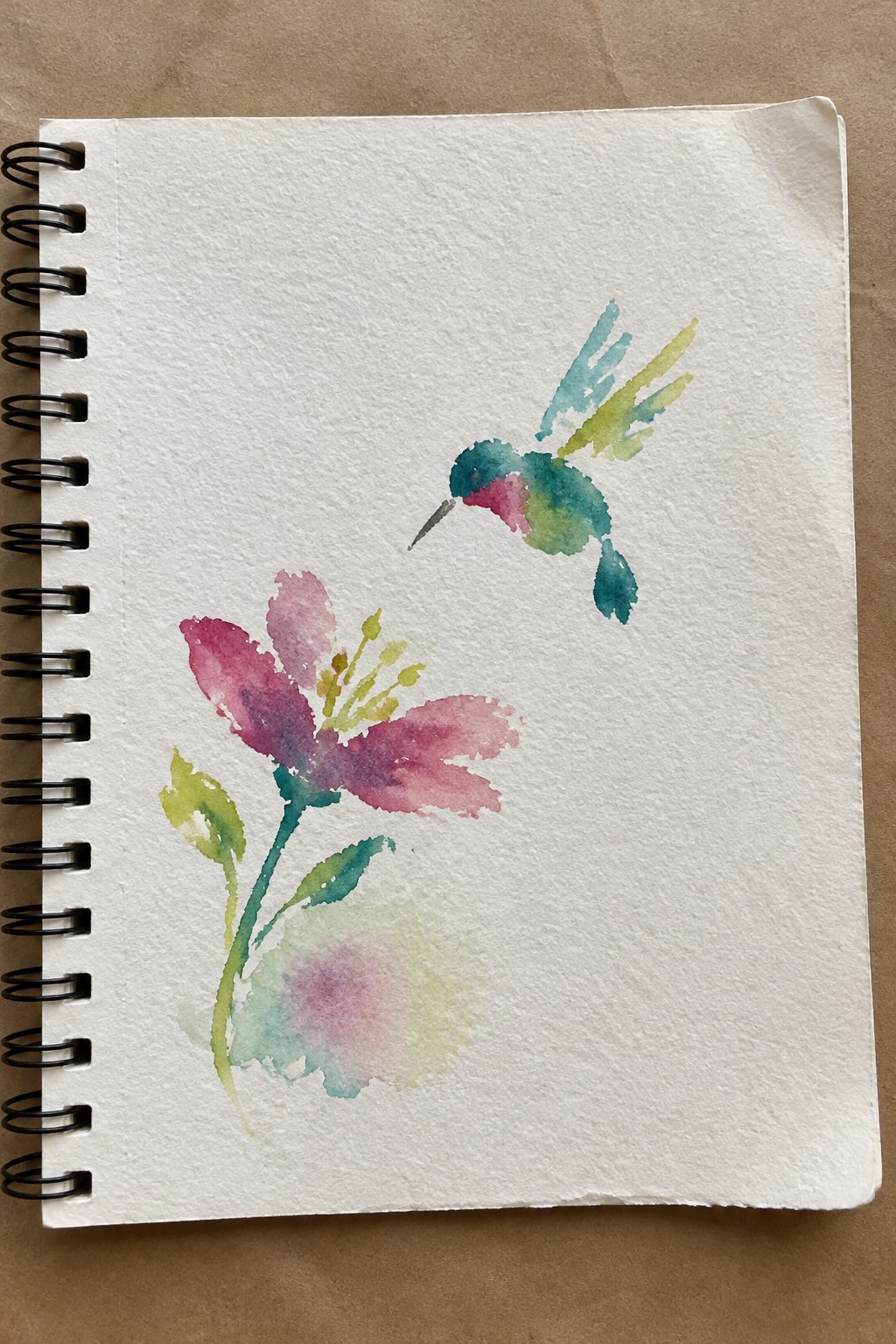 Watercolor hummingbird in teal and pink hovering above a magenta flower in a sketchbook.