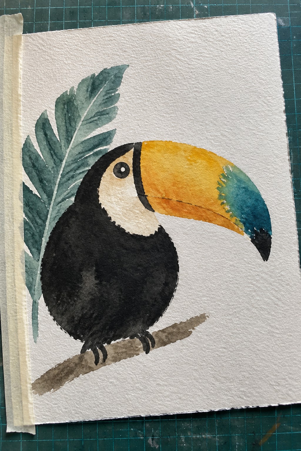 A watercolor painting of a toucan with a black body, white face, and yellow-blue beak perched on a branch next to a green leaf.