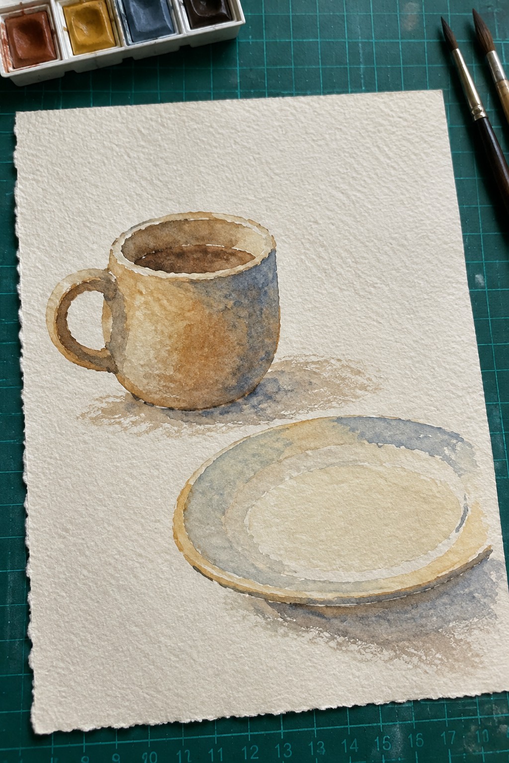 Watercolor painting of brown-blue cup and saucer beside palette and brushes on paper