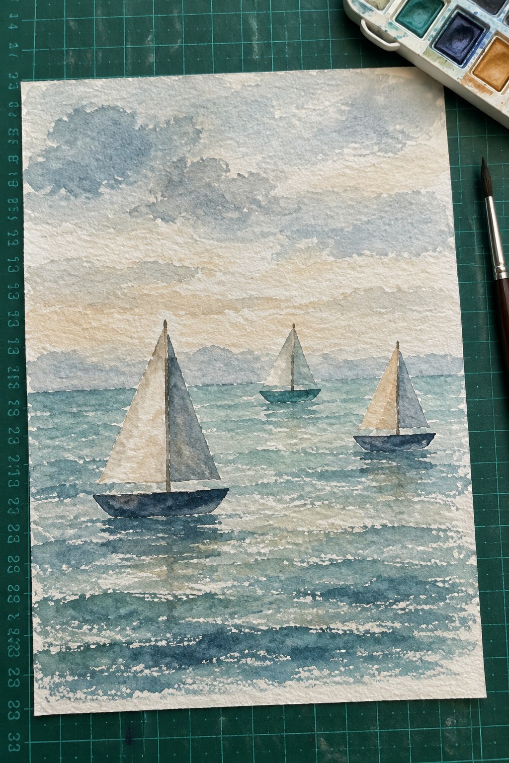 Watercolor seascape of three sailboats on blue wavy sea, palette and brush on green mat.