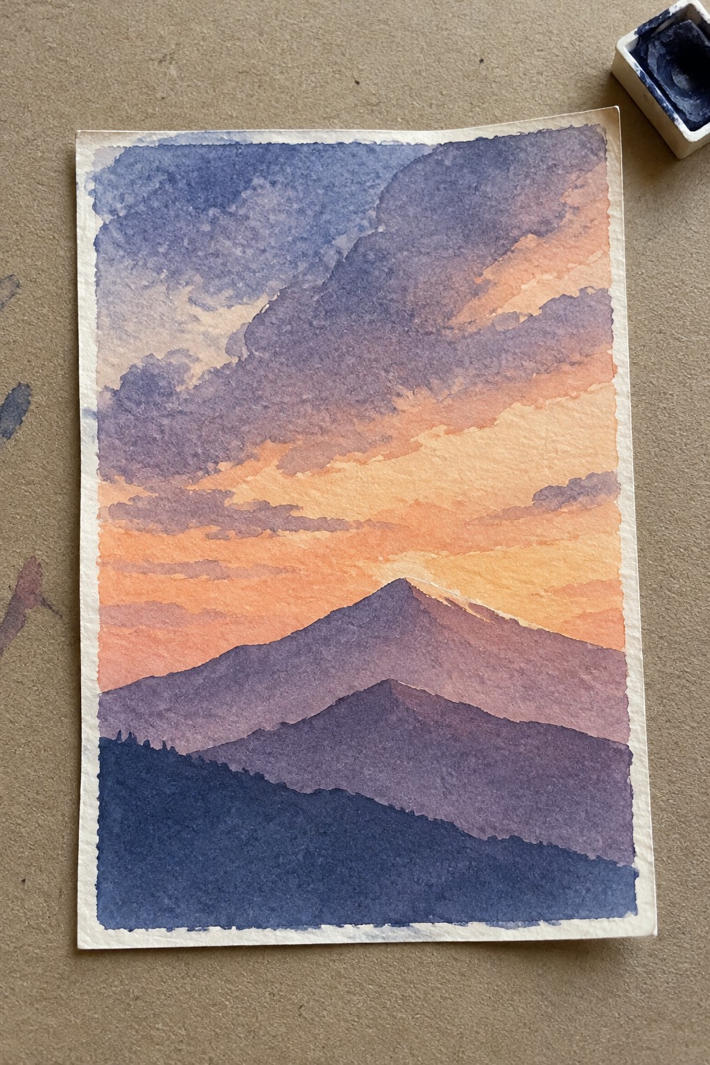 Watercolor of layered purple mountains under orange-peach sunset sky with ink pan nearby