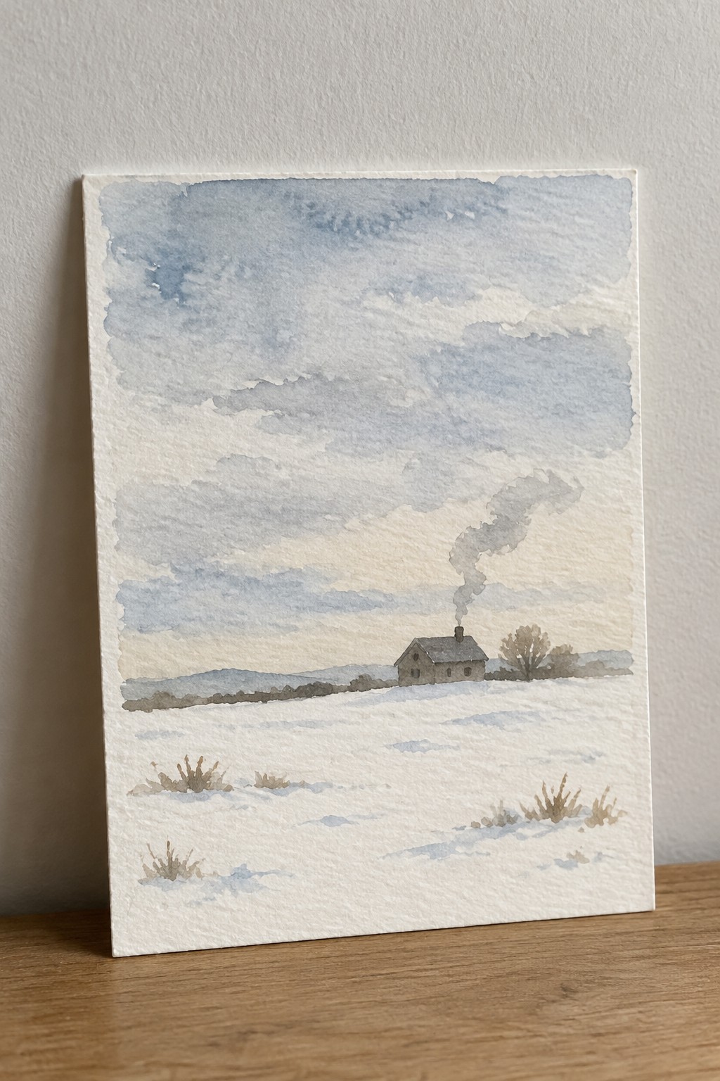 Watercolor of snowy field with house emitting smoke, bare shrubs, and distant hills under cloudy sky.
