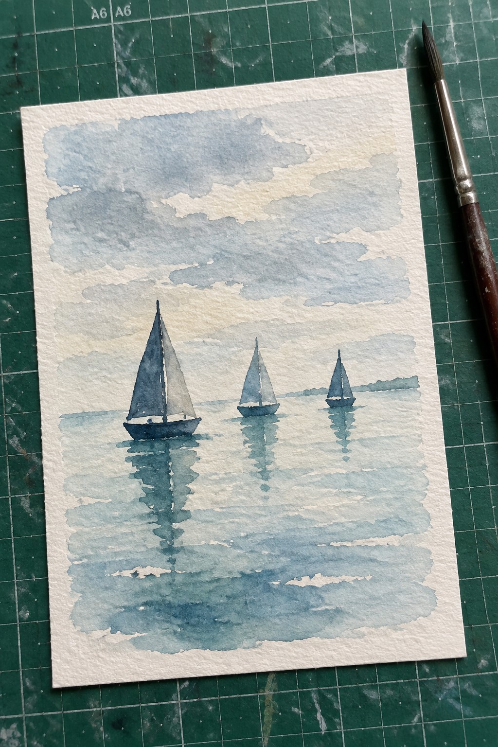 Watercolor of three sailboats with reflections on blue water beside paintbrush on mat.