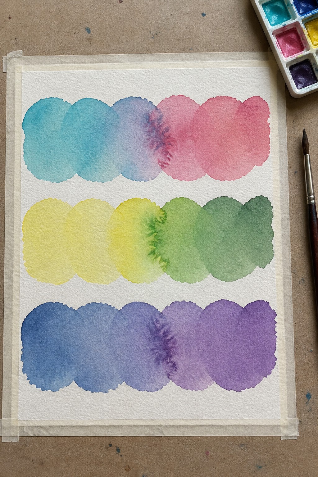 Watercolor paper with blue-pink, yellow-green, blue-purple gradients, palette and brush nearby