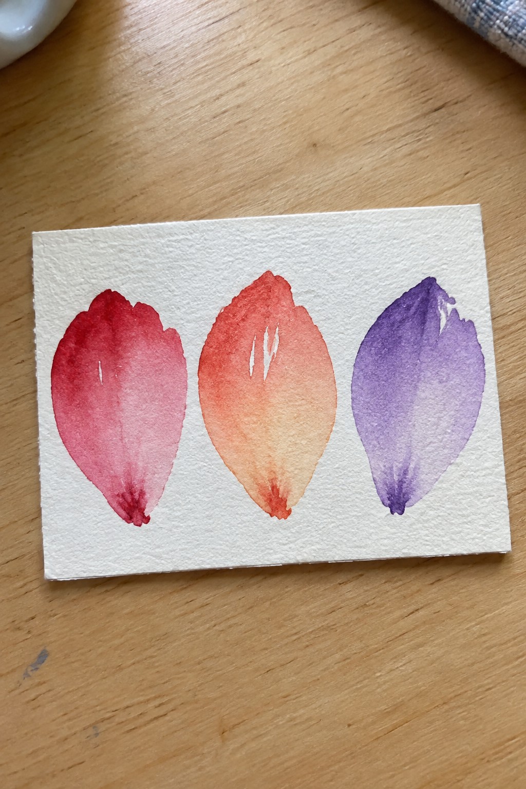 Three gradient watercolor petals in pink, orange, and purple on white paper over wood