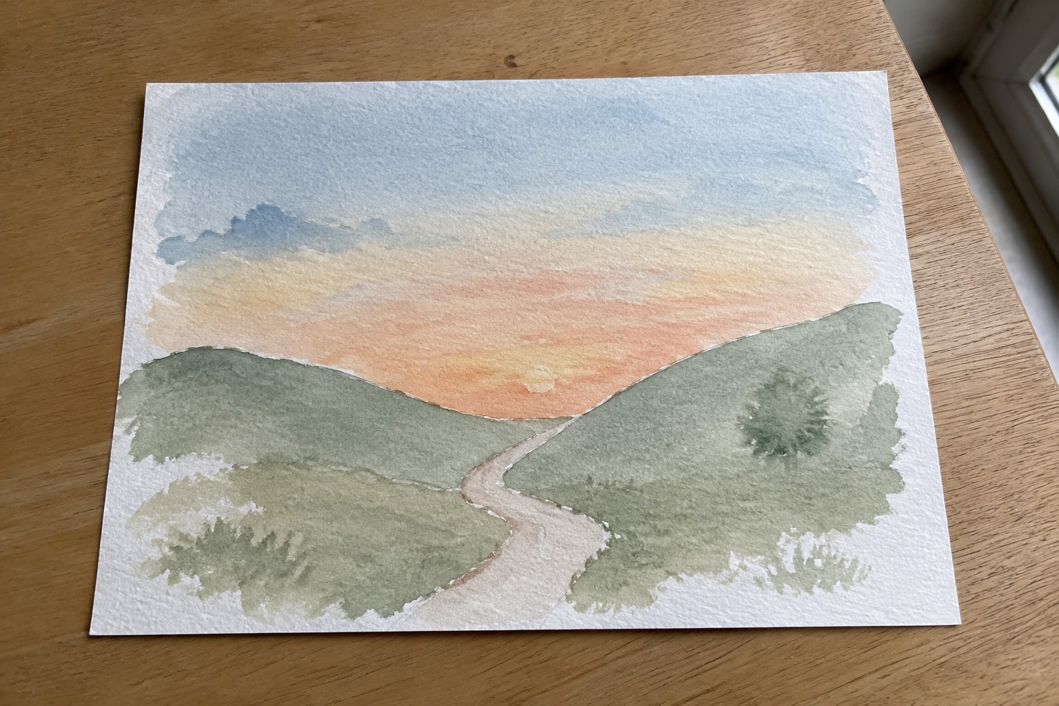 Watercolor of winding path through green hills under orange-pink sunset sky