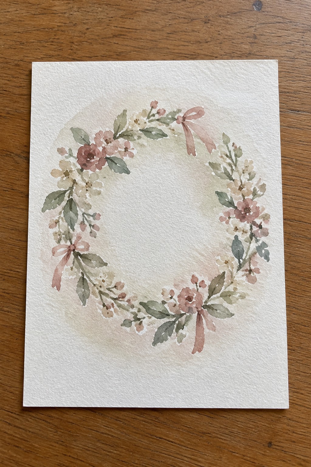 Watercolor floral wreath with pink blooms, green leaves, and ribbon bows on white paper over wood