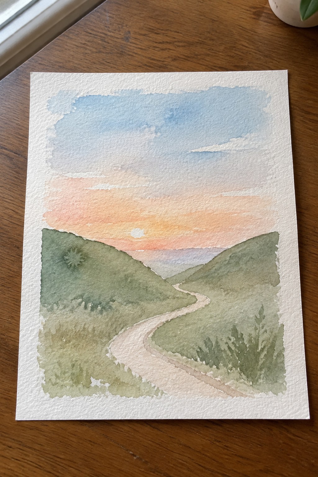 Watercolor of winding path through green hills under orange-pink sunset sky