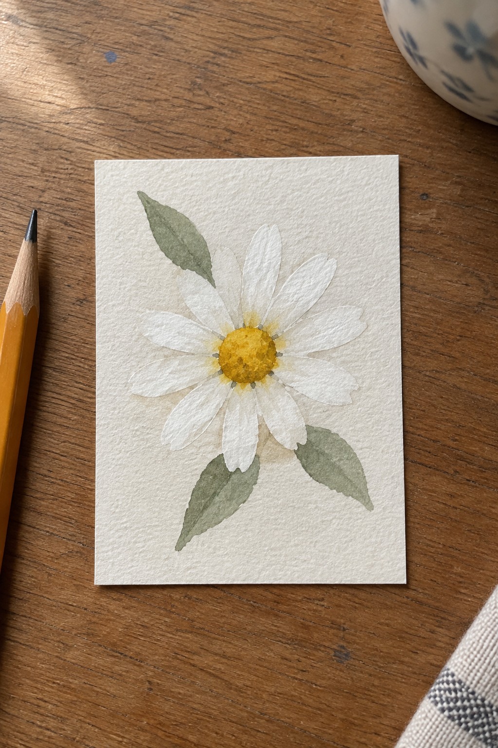 Watercolor daisy with yellow center and green leaves on paper beside pencil on wood table