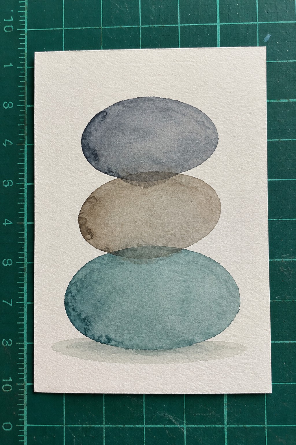 Watercolor of three stacked ovals in gray, beige, and teal with shadow on white paper