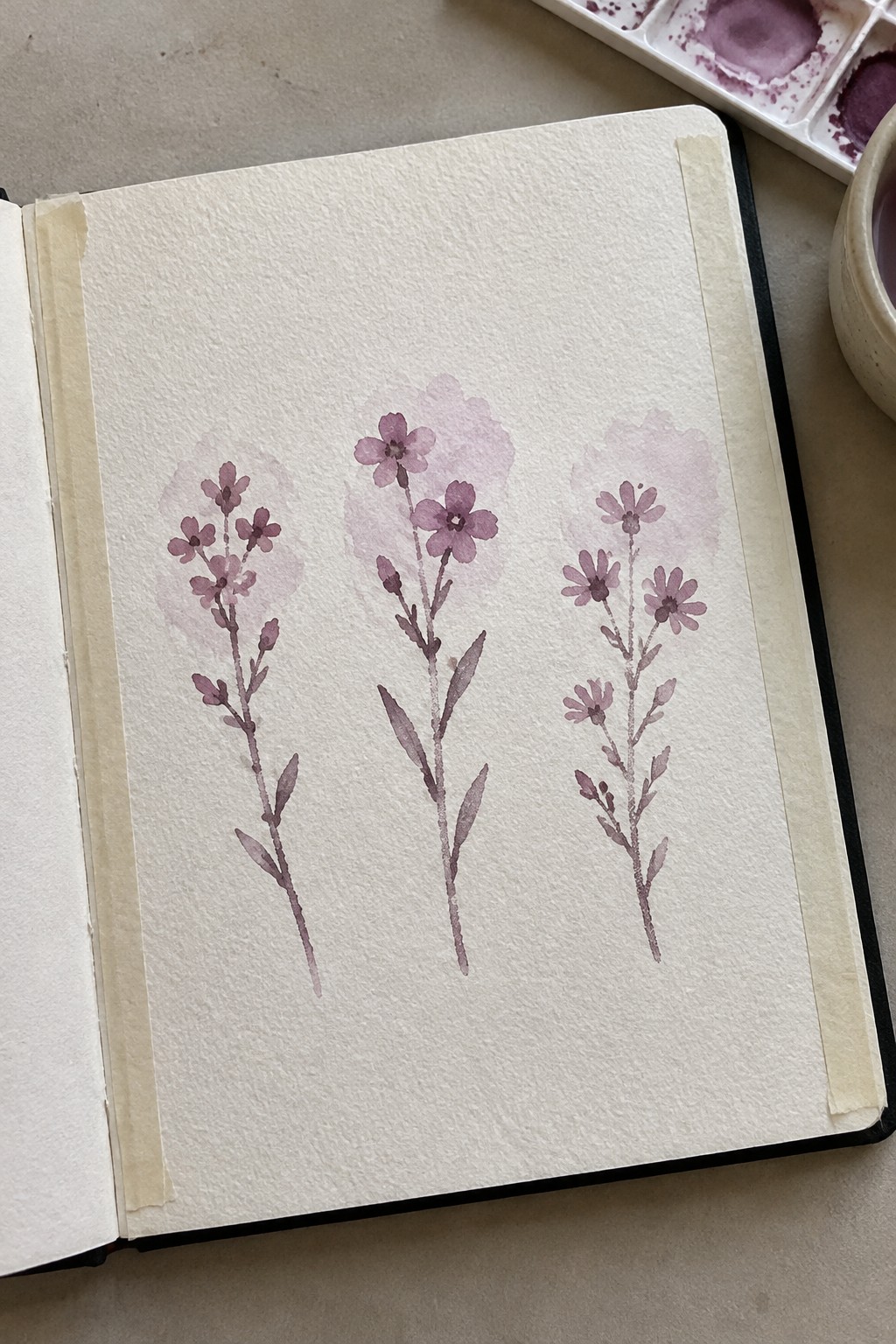 Open sketchbook with three purple watercolor flower paintings and visible paint palette