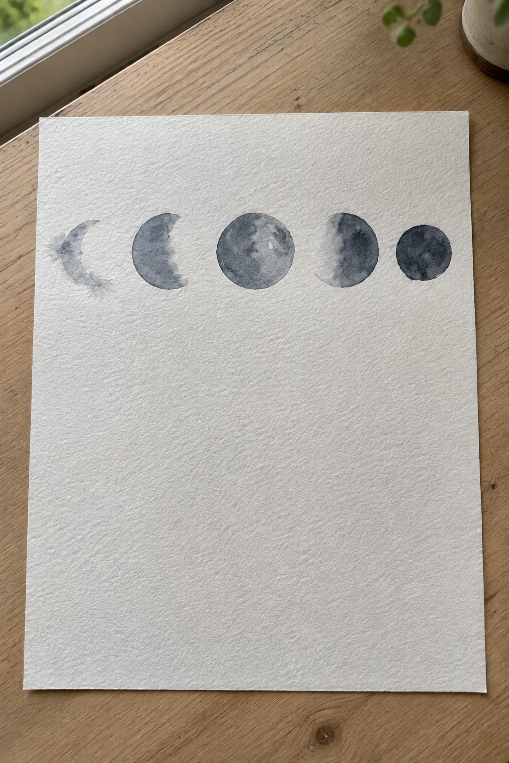 Watercolor moon phases in gray tones on white paper over wooden table