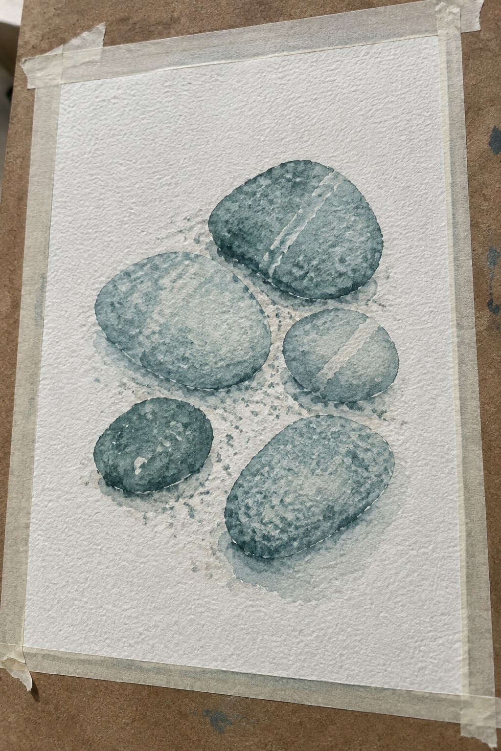 Five teal watercolor pebbles on textured white paper taped to brown surface