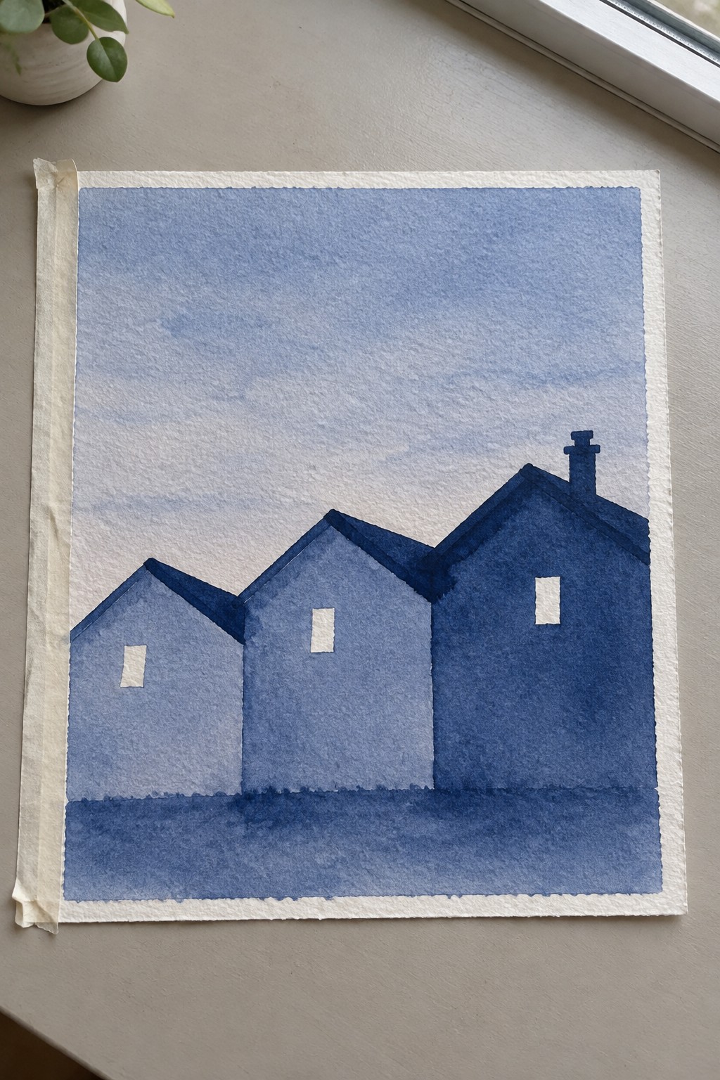 Watercolor of three blue houses with white windows against gradient sky on taped paper
