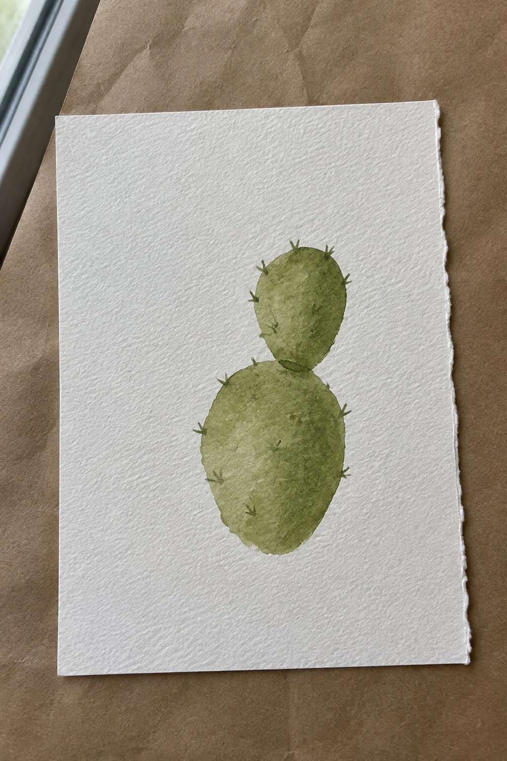 Watercolor painting of two-lobed green cactus with spines on white paper over brown surface.