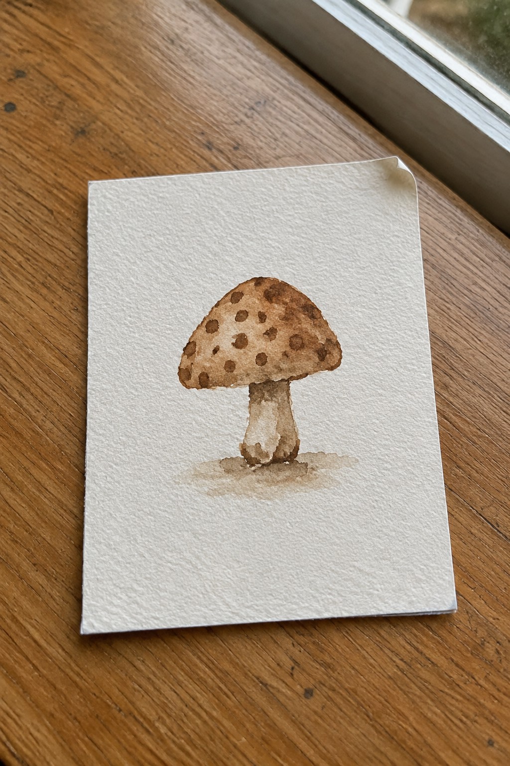 Watercolor of brown spotted mushroom on white paper over wooden windowsill