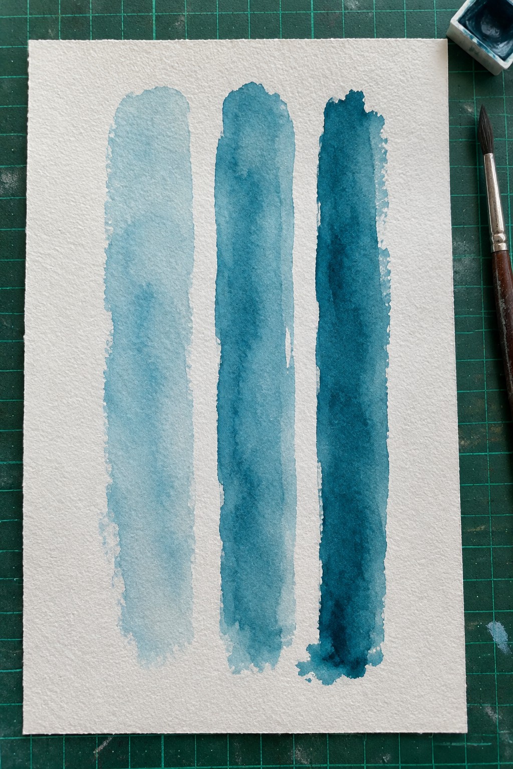 Three vertical blue watercolor strokes on white paper beside paintbrush on green mat