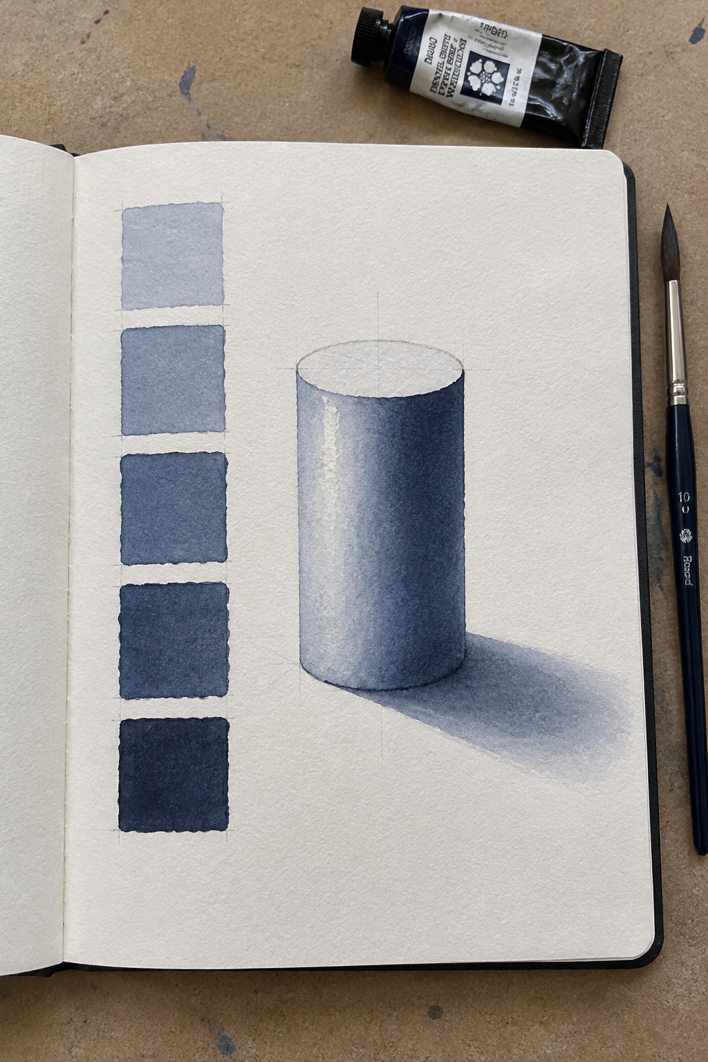Open sketchbook with blue watercolor swatches and shaded cylinder, paint tube and brush nearby.