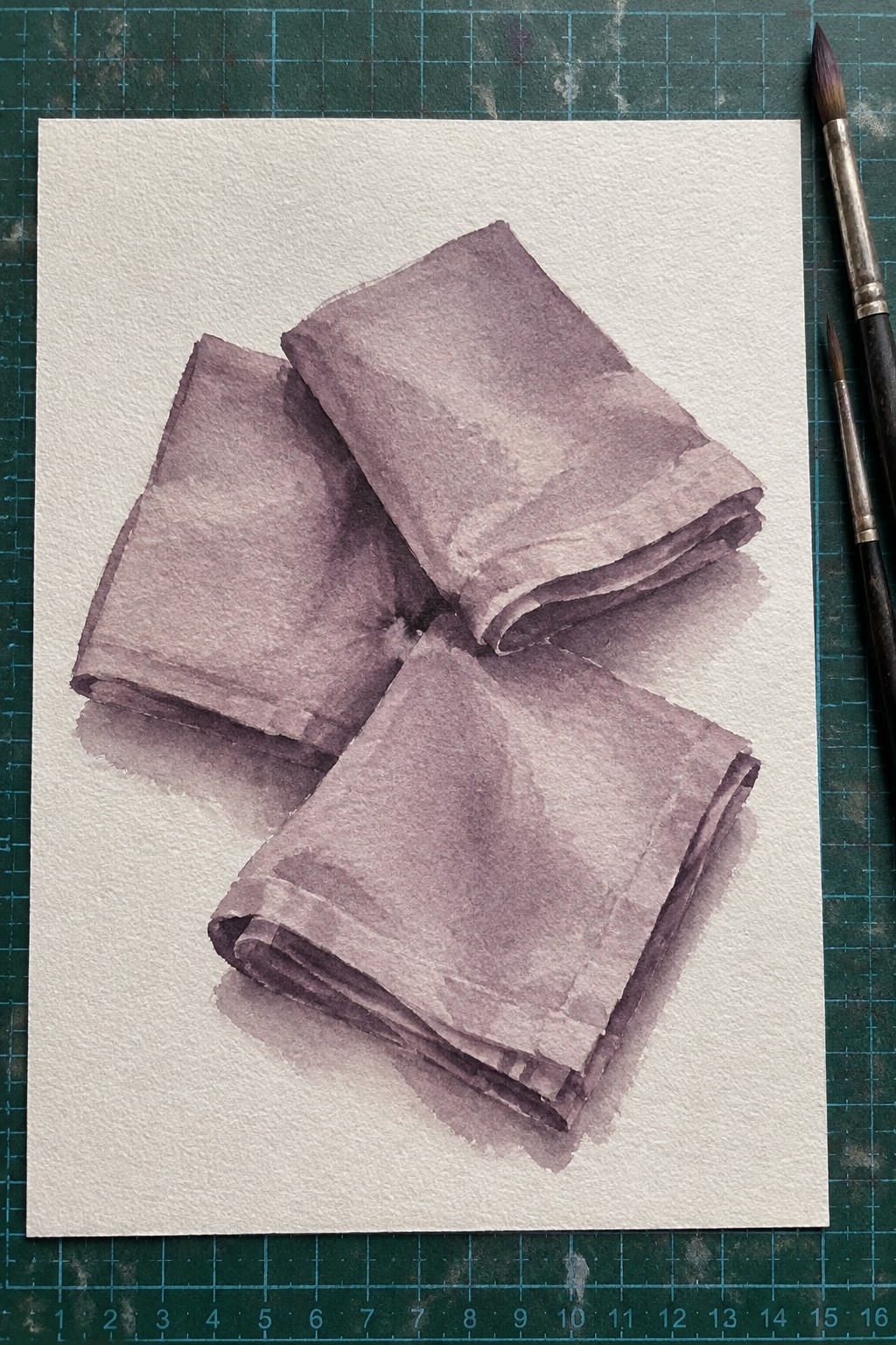 Watercolor painting of three folded purple cloths beside paintbrushes on white paper
