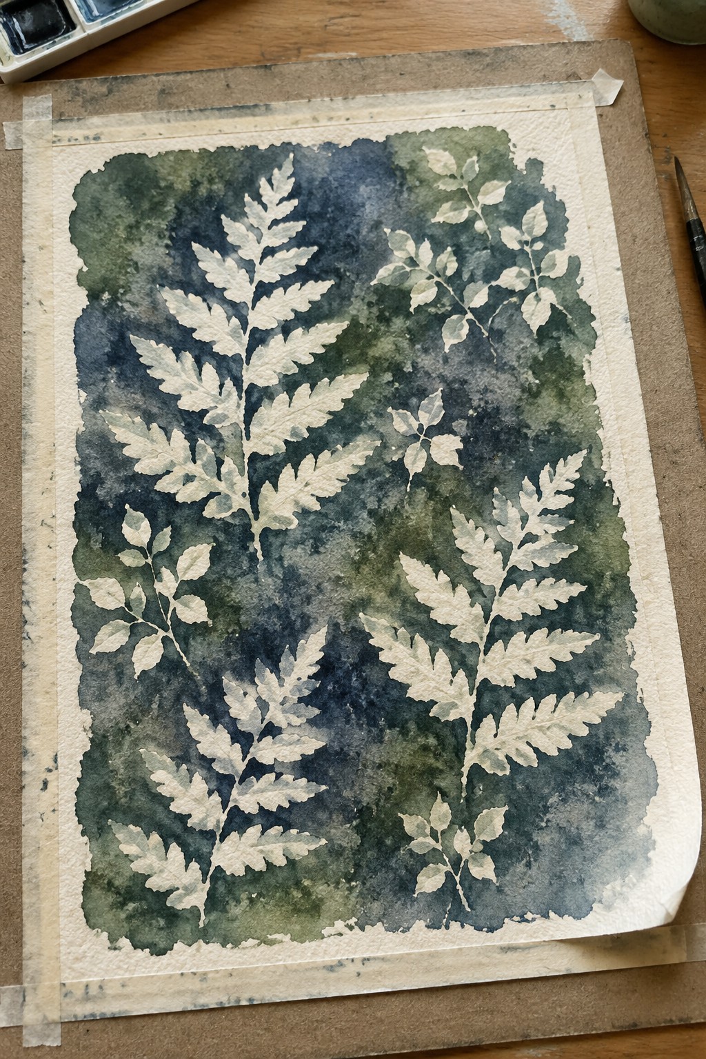 Pale fern leaves in negative space on dark blue-green watercolor, taped on wooden table.