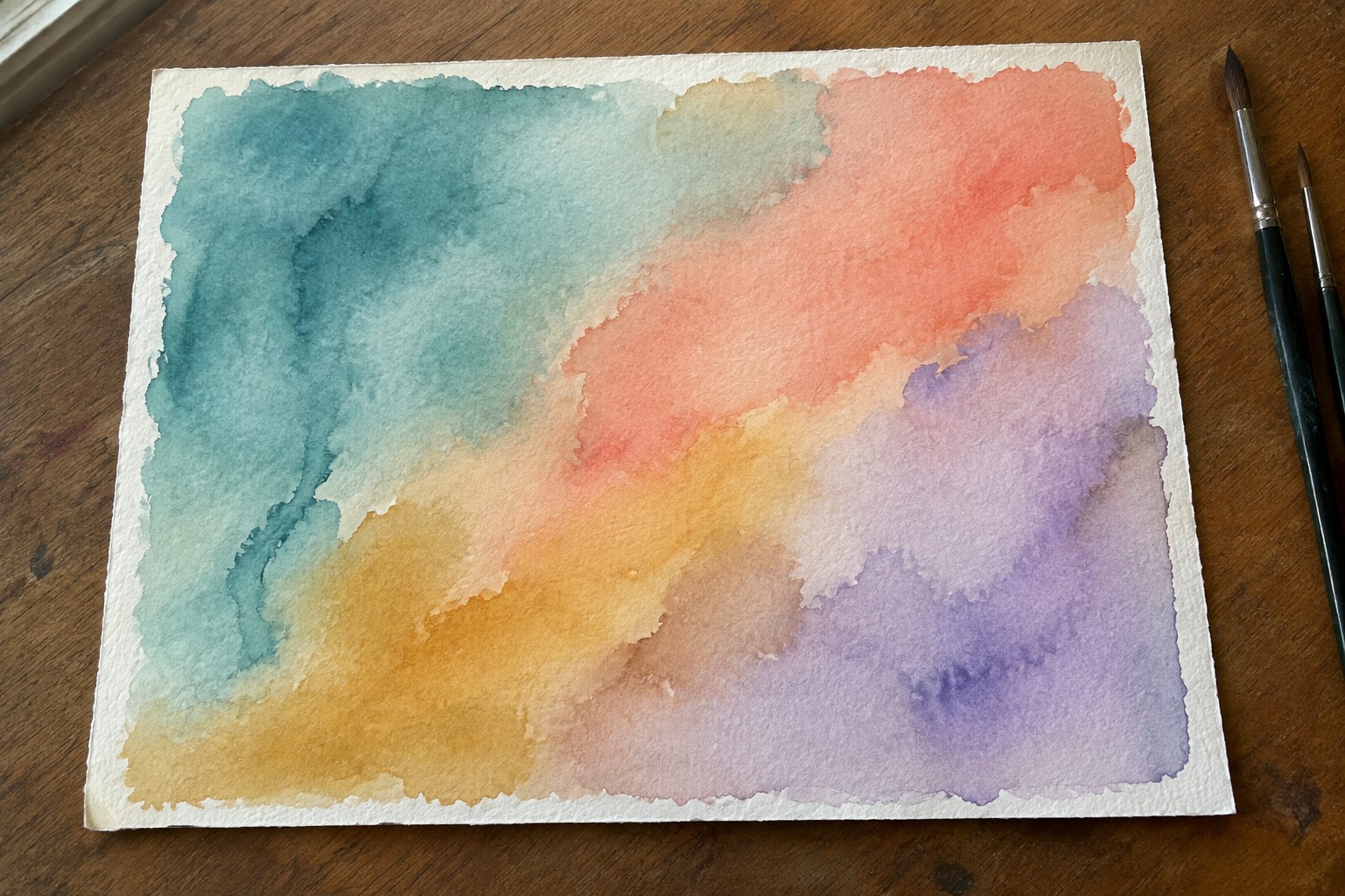 A rectangular sheet of watercolor paper on a wooden table shows abstract blended washes of teal, coral, yellow, and purple with soft edges and two paintbrushes resting to the right.