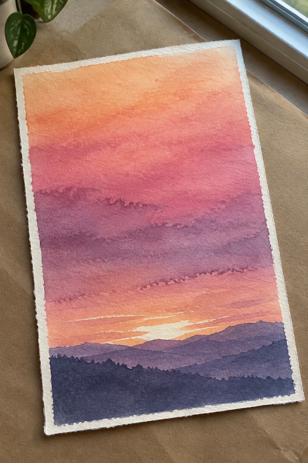 Watercolor sunset with orange-pink-purple sky gradients over layered dark mountain silhouettes on textured paper.