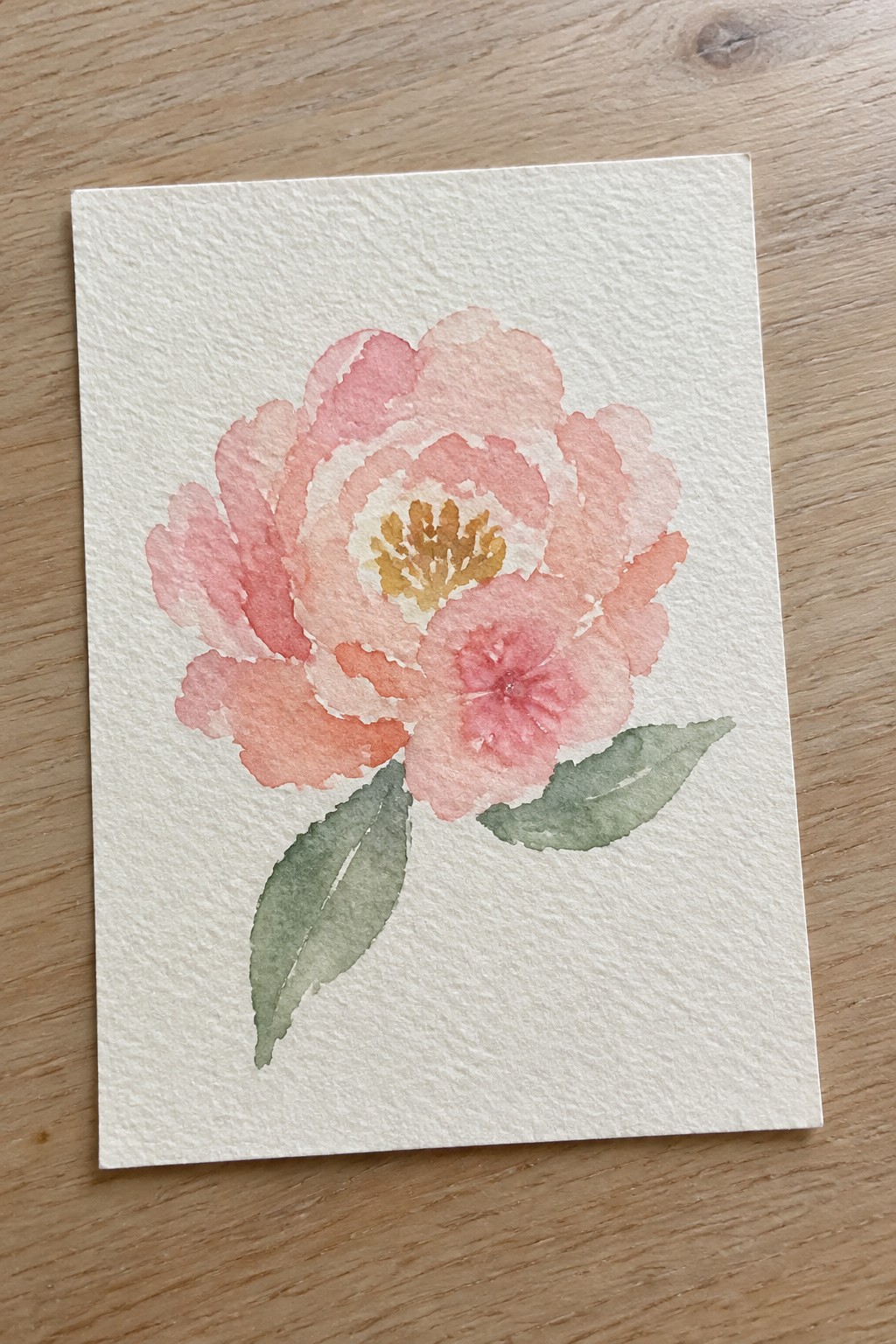 Watercolor painting of pink peony with yellow center and green leaves on white paper