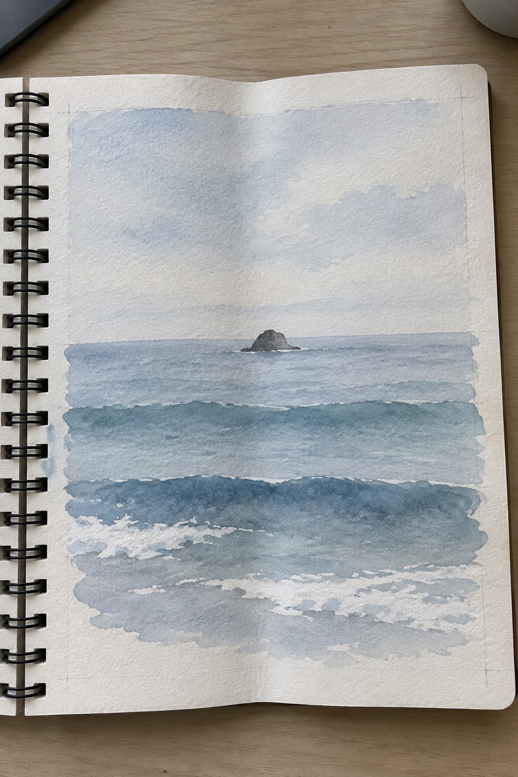 Watercolor seascape in open sketchbook showing layered blue waves and distant rock