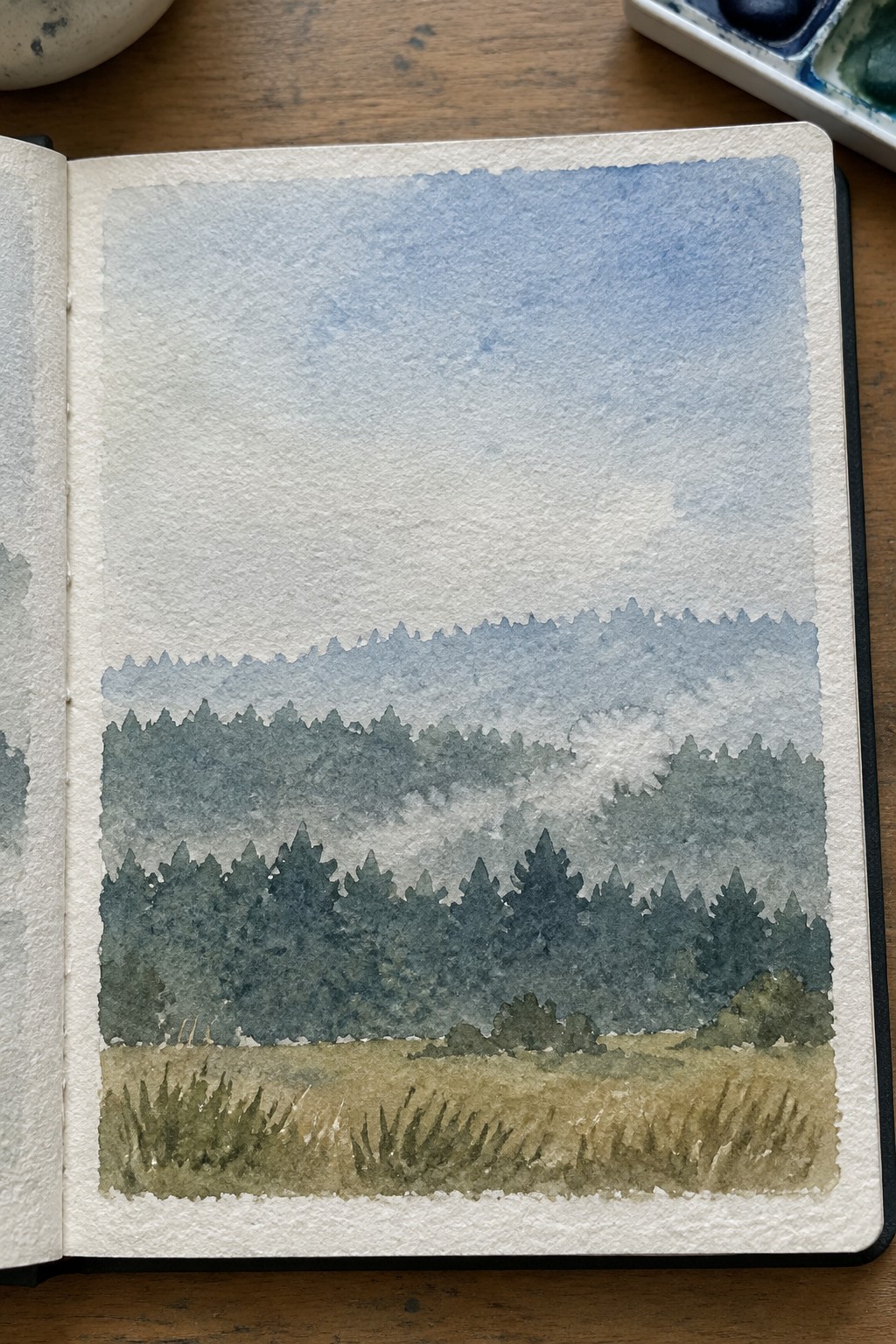 Open sketchbook with watercolor of misty layered green forests and blue sky