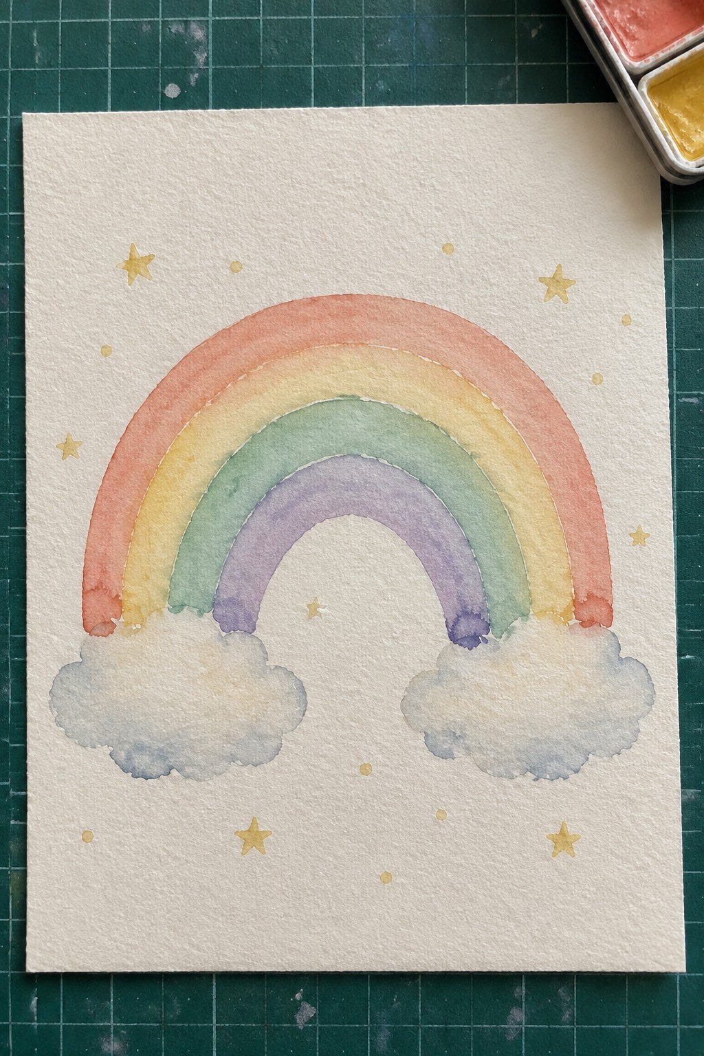 A watercolor rainbow with blended pastel bands, two clouds at the base, and small gold stars scattered on white paper.