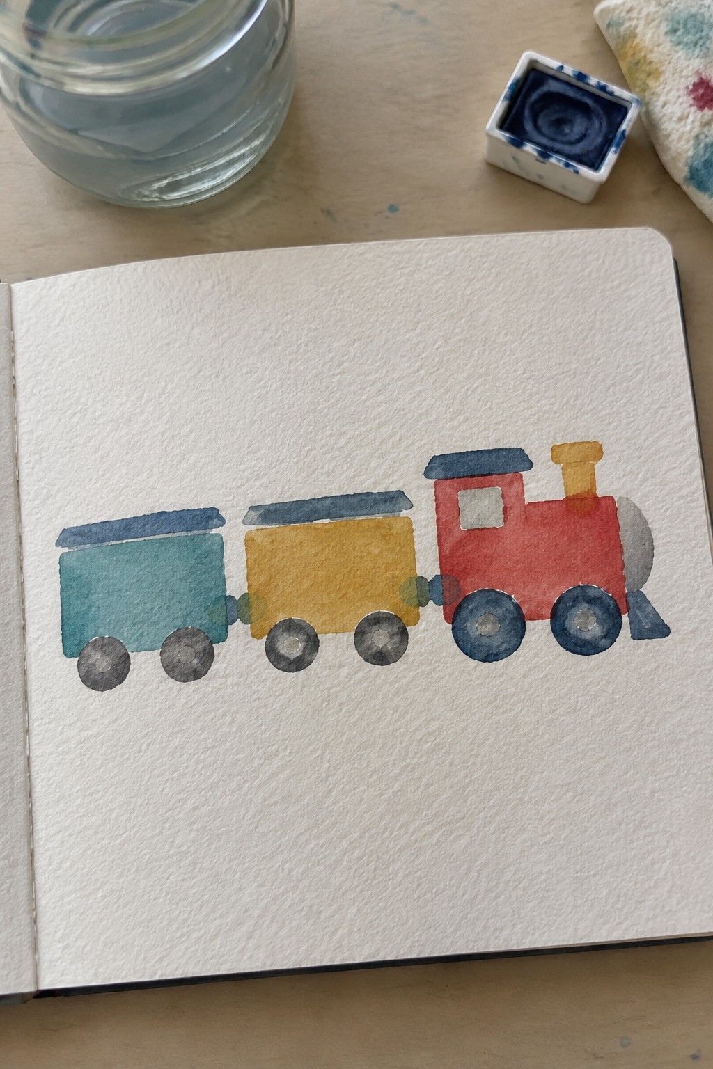 Open sketchbook with watercolor train in red, yellow, and teal beside paint supplies