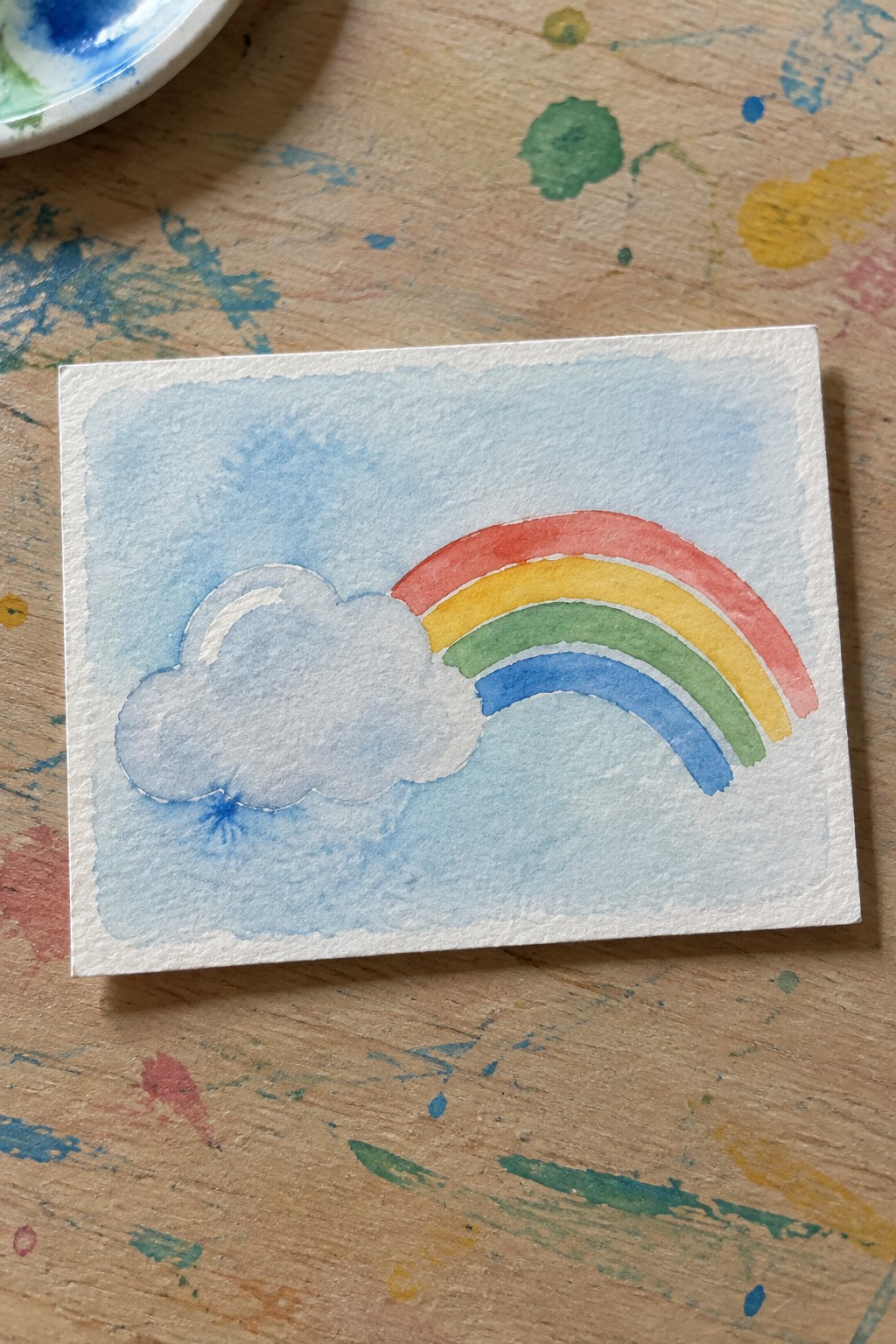 Watercolor rainbow arching from a cloud on paint-splattered wood table