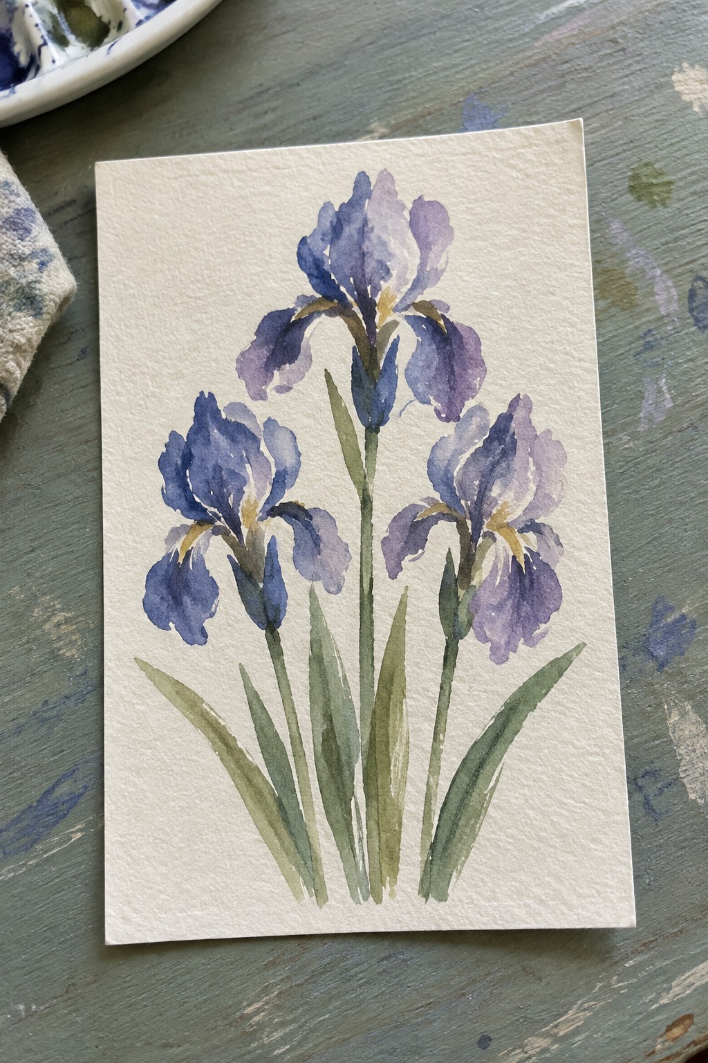 Watercolor painting of three purple irises with yellow accents and green leaves on paper