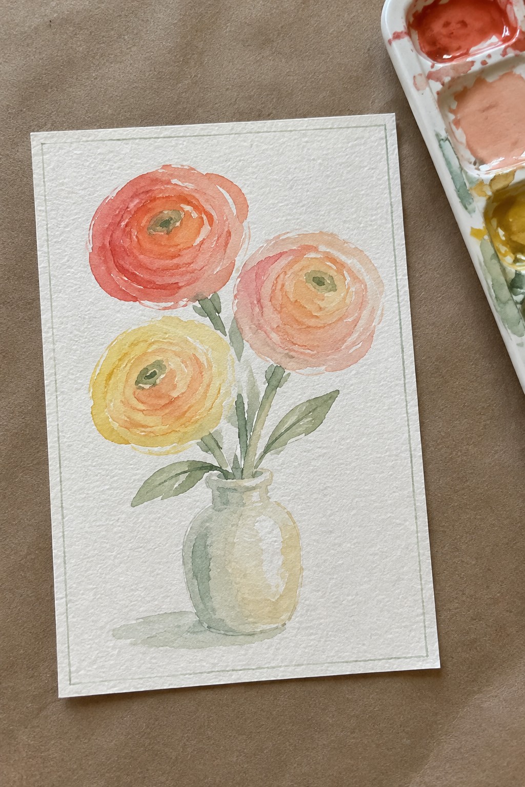 Watercolor painting of three colorful flowers in a vase beside paint palette