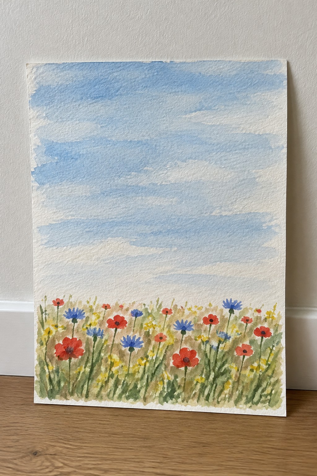 Watercolor of red poppies and blue cornflowers blooming in a grassy meadow under blue sky