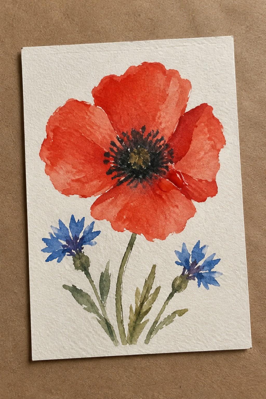 A watercolor painting of a large red poppy with a dark center above two small blue cornflowers on white paper.