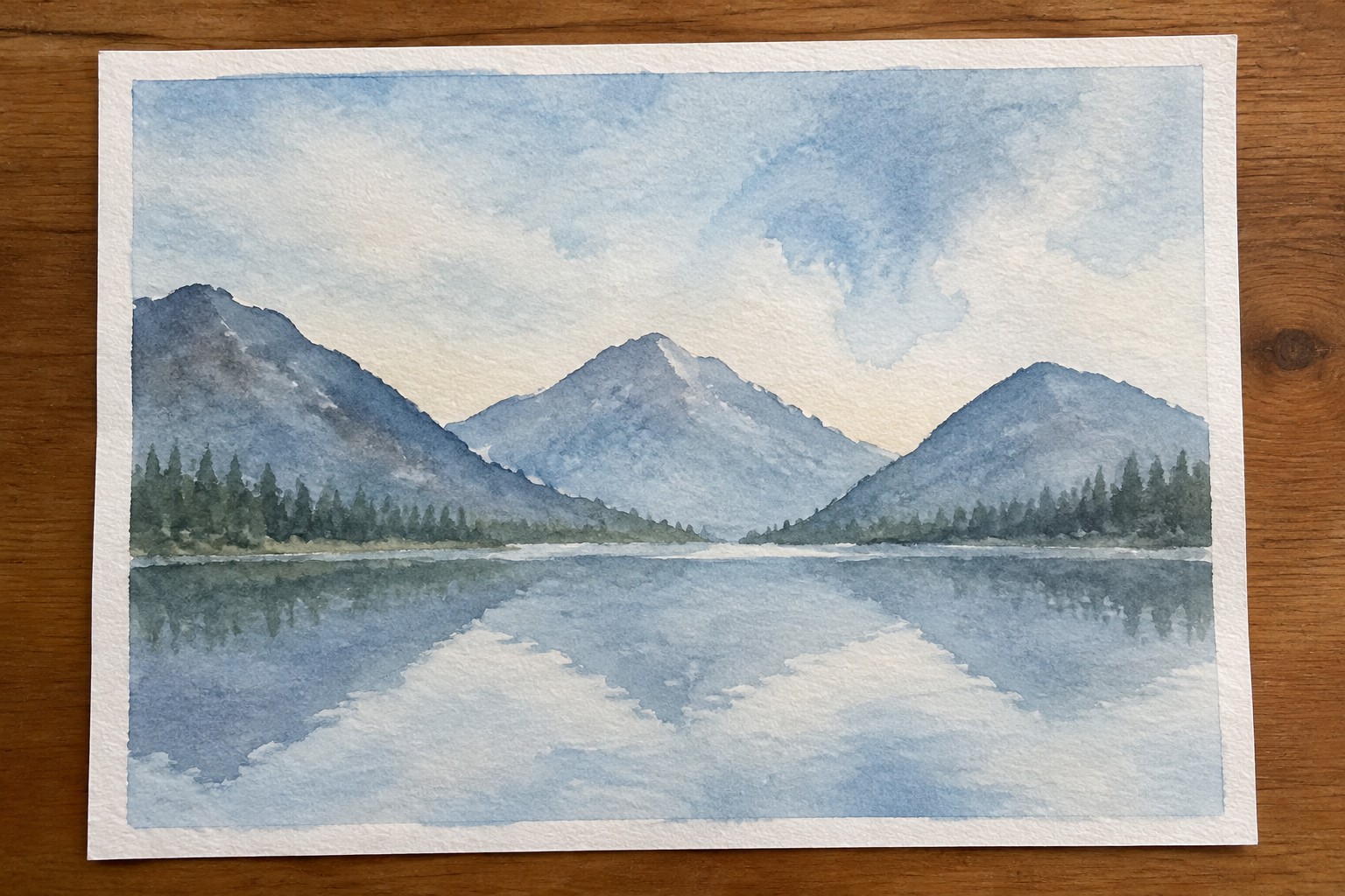 Watercolor of blue mountains reflected in calm lake with pine trees under cloudy sky.
