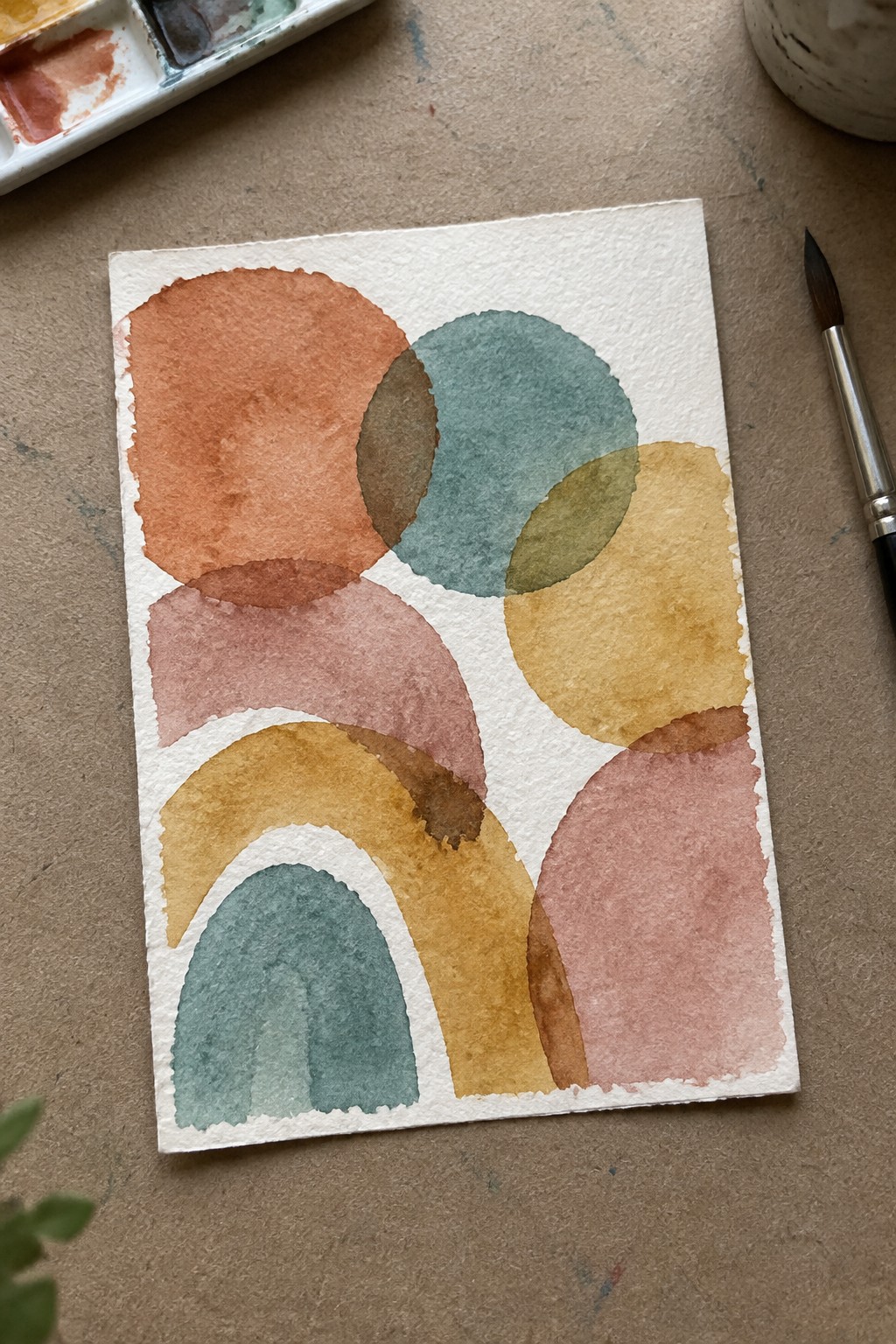 Abstract watercolor with overlapping circles and arches in terracotta, teal, and mustard beside brush and palette