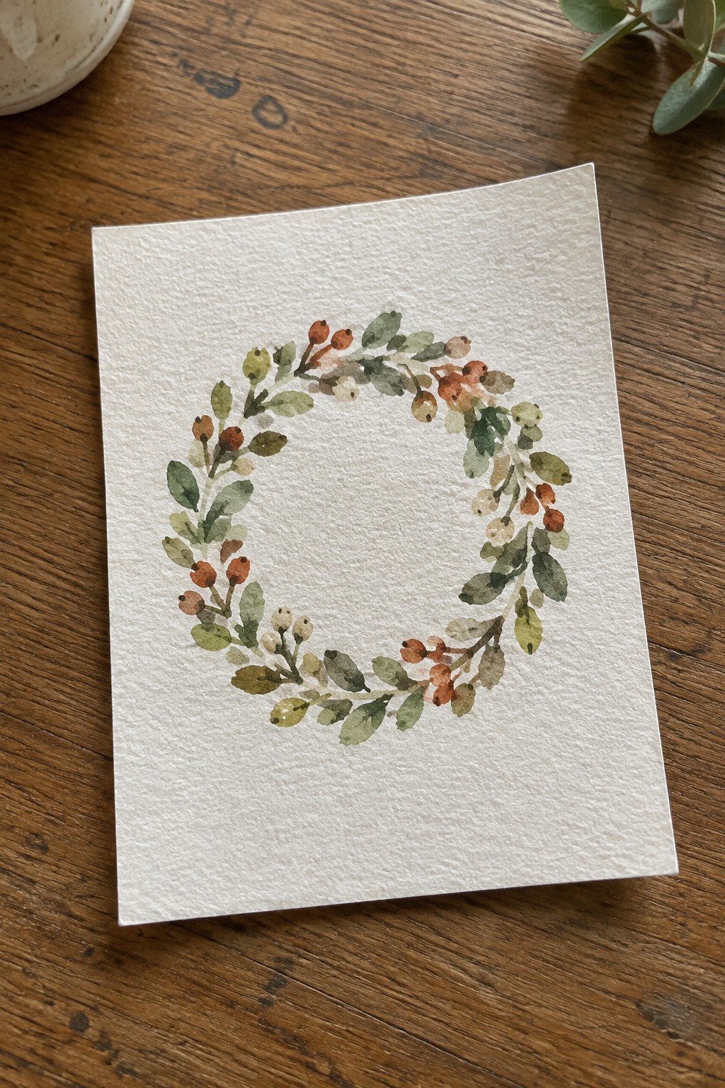 Watercolor painting of a circular wreath made of green leaves and orange berries on textured white paper resting on wood.