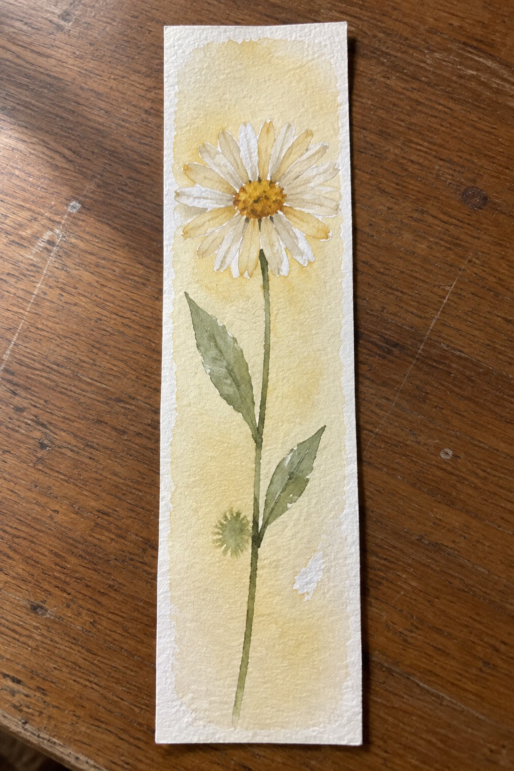Watercolor bookmark with a daisy flower, stem, two leaves, and small bud on a yellow gradient background.