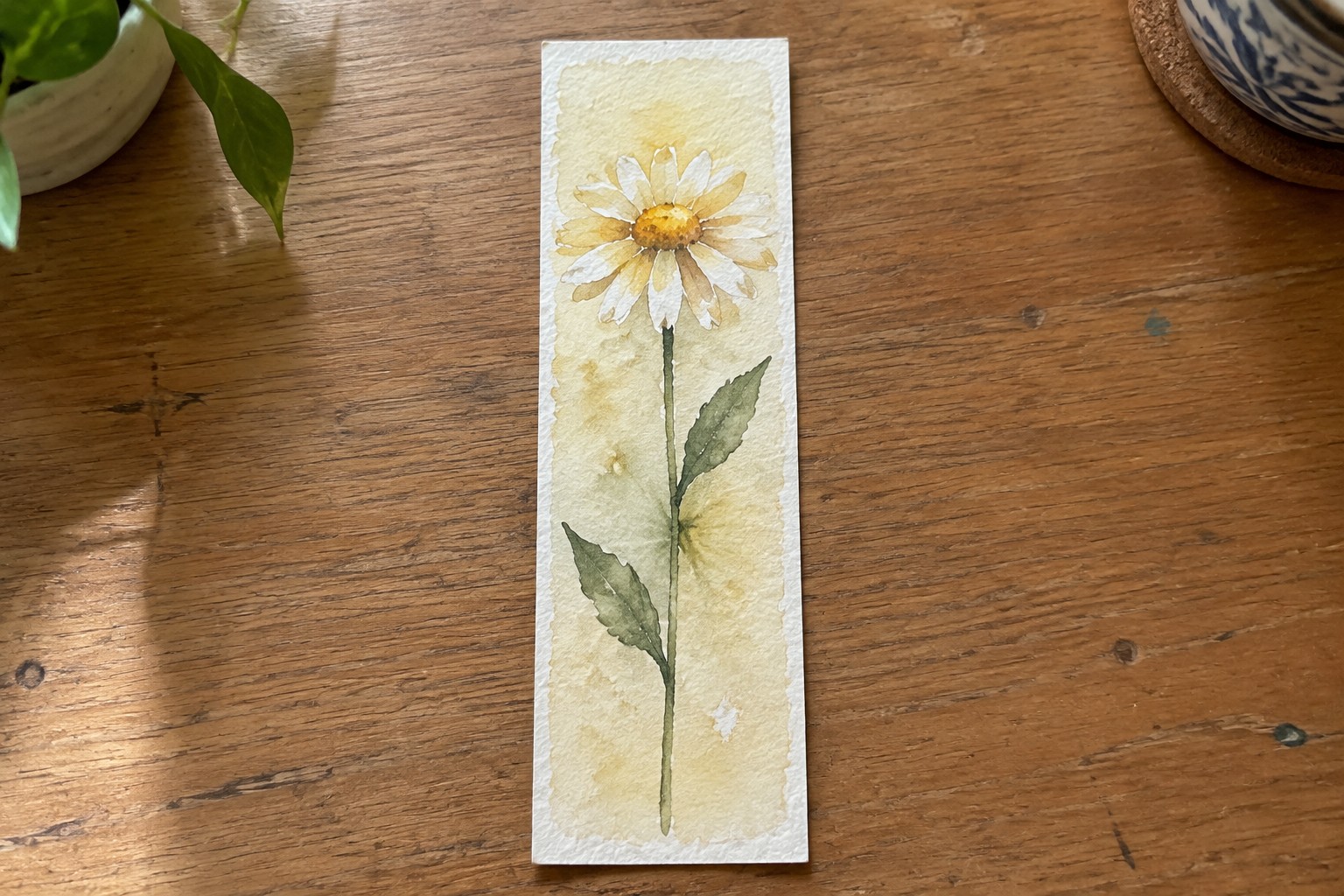 Watercolor bookmark with a daisy flower, stem, two leaves, and small bud on a yellow gradient background.