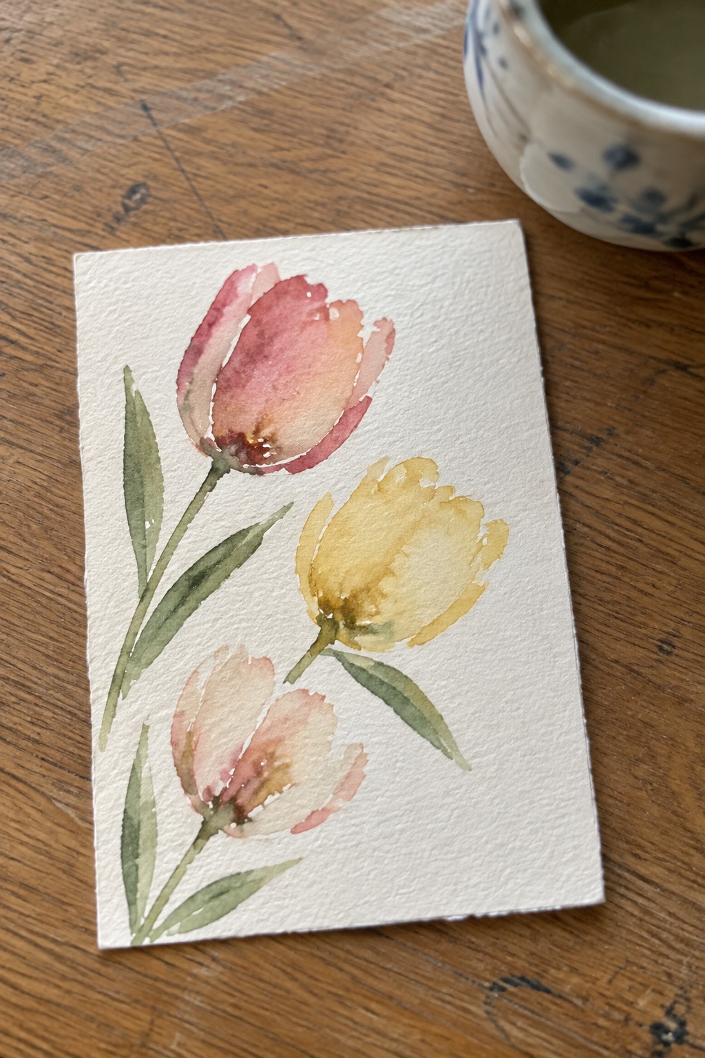 Watercolor tulips in pink, peach, and yellow with green leaves on white paper