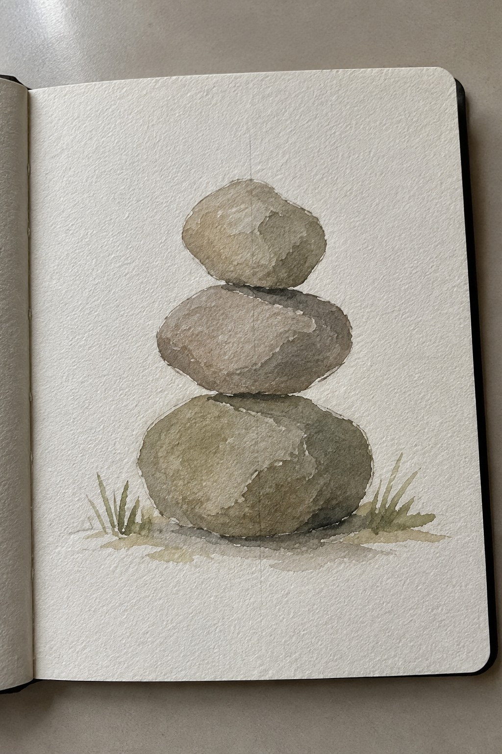 Watercolor painting of three stacked stones with grass in an open sketchbook