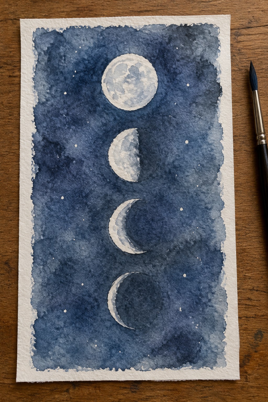 Watercolor painting of four moon phases stacked vertically on dark blue starry wash with paintbrush on wooden table.