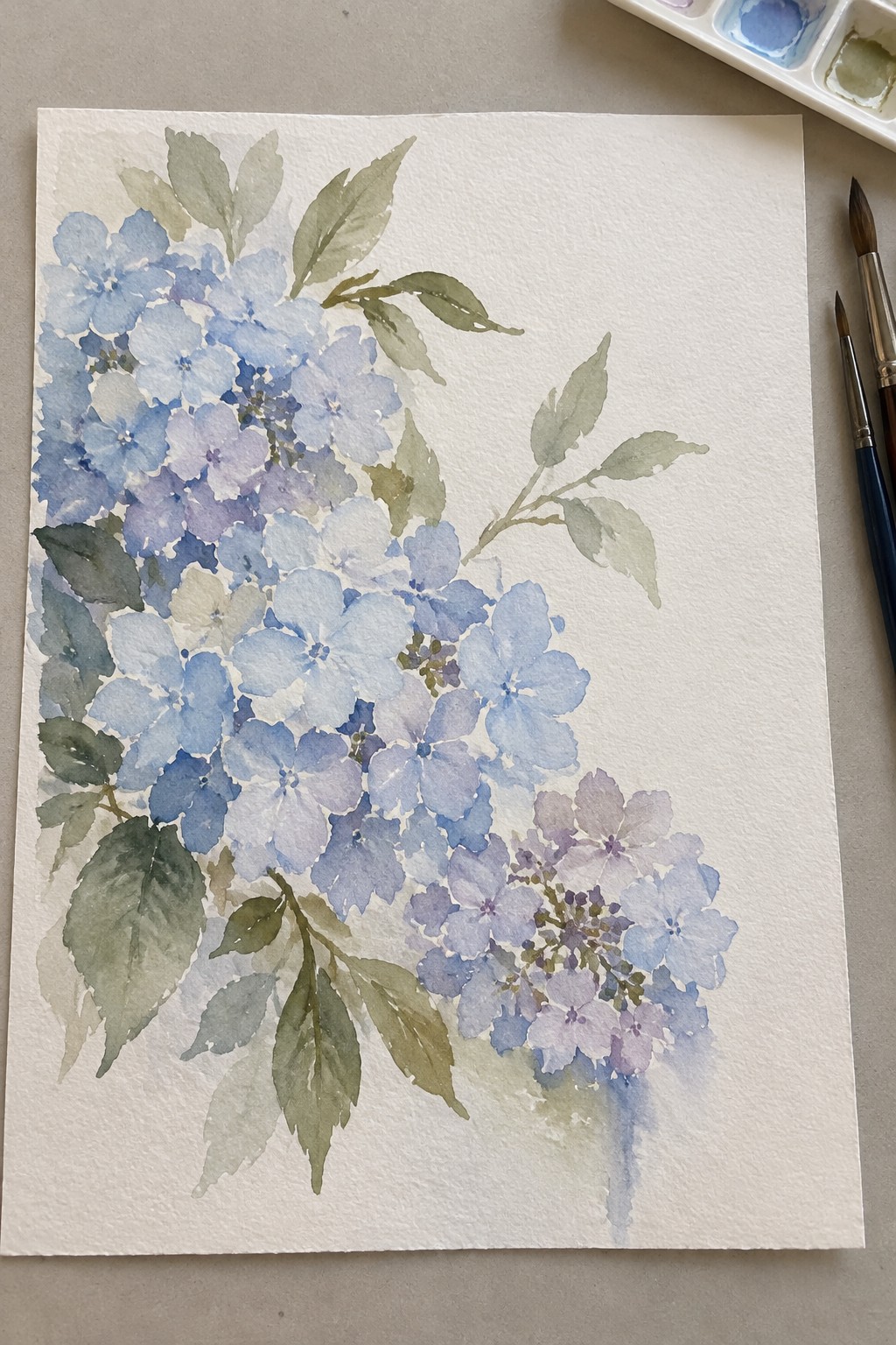 Watercolor painting of blue hydrangeas with green leaves, brushes, and palette