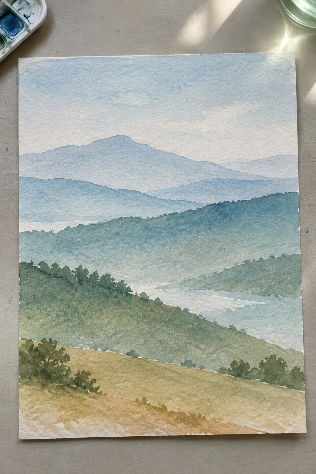 Watercolor landscape of layered blue mountains, misty green hills, and golden foreground field.